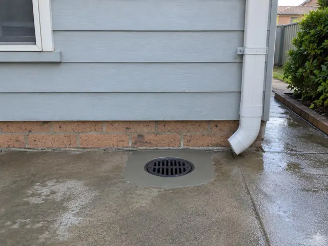 Clean outdoor drain grate beside wet concrete paving and house wall in Minchinbury