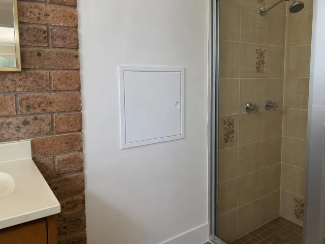 Small access panel in a painted bathroom wall beside tiled shower and brick feature wall in Mount Druitt